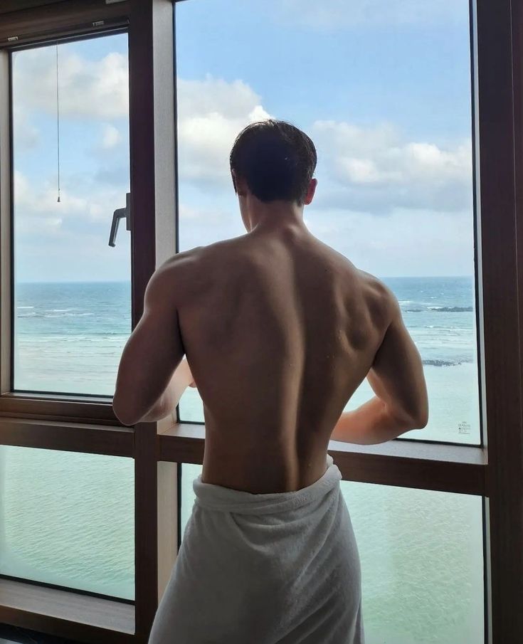 a man standing in front of a window looking out at the ocean with his back to the camera