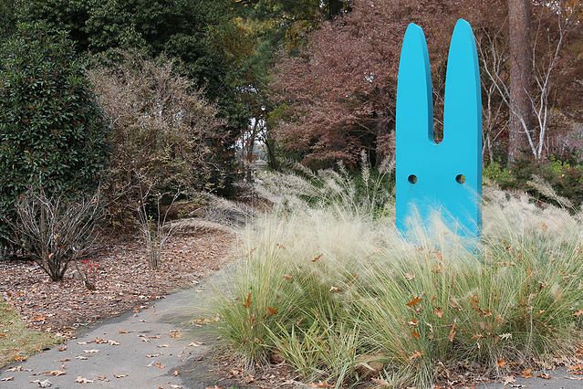 a blue bunny statue sitting in the middle of a park next to tall grass and trees