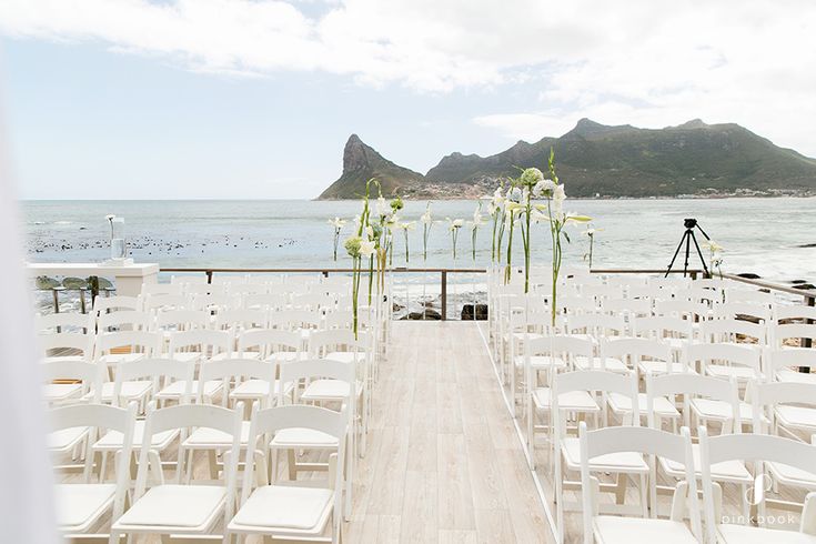 an outdoor ceremony set up with white chairs and flowers on the aisle, overlooking the ocean