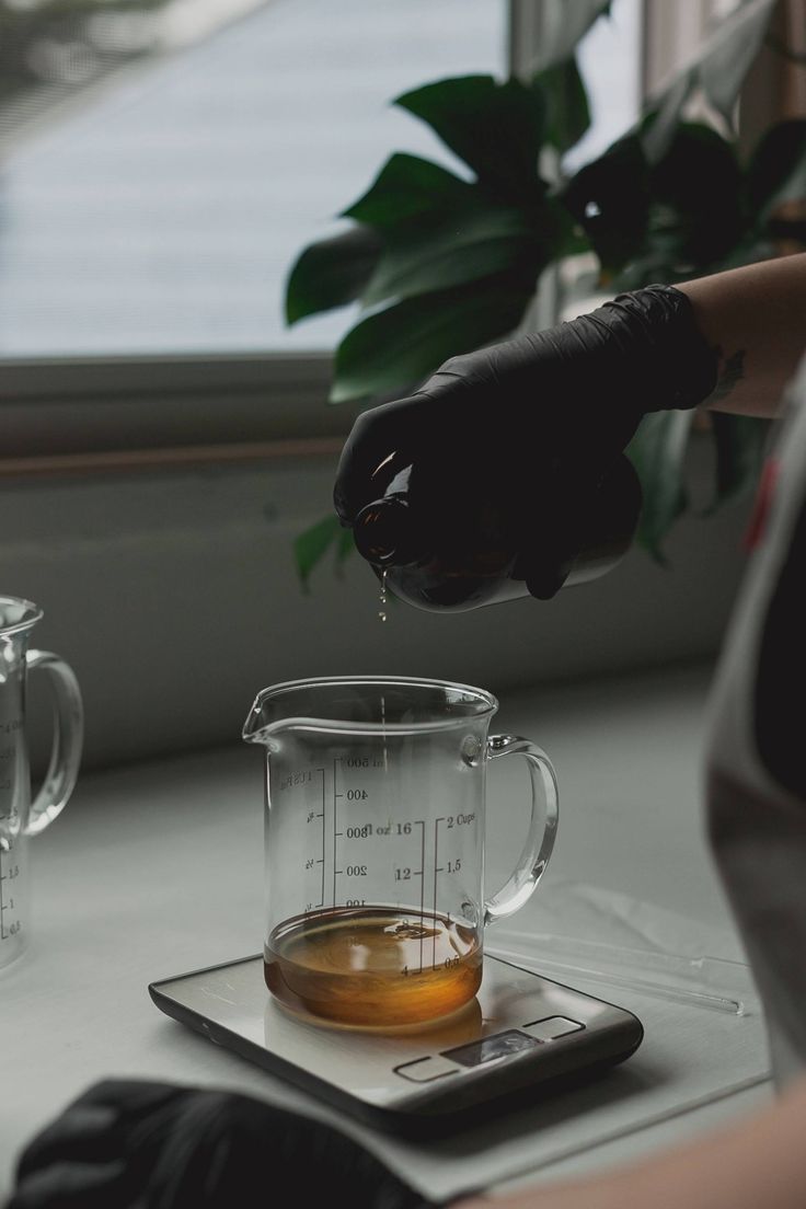 a person pouring tea into a glass pitcher on top of a scale next to a potted plant