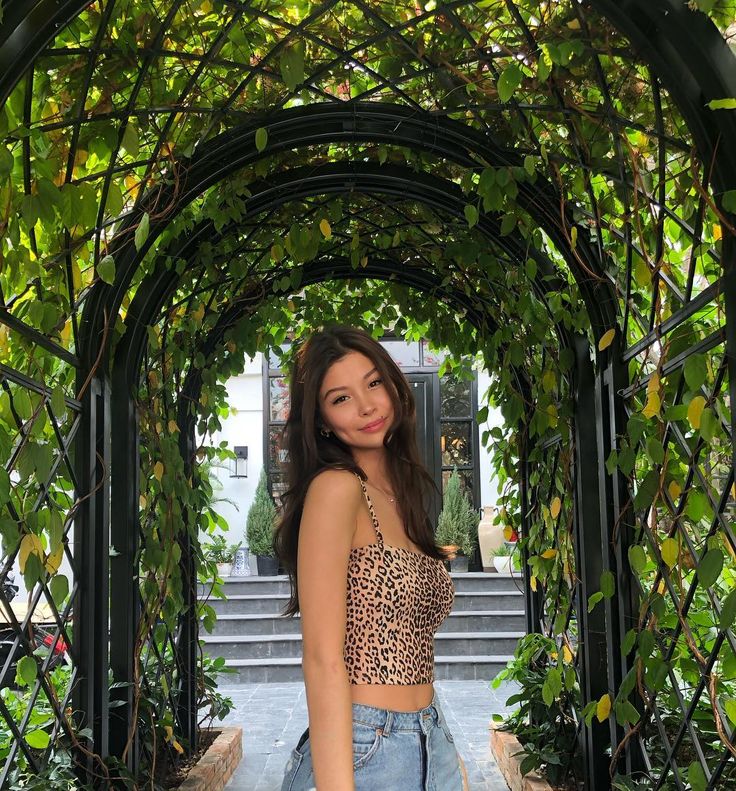 a beautiful young woman standing under an arch covered in green leaves and vines, smiling at the camera