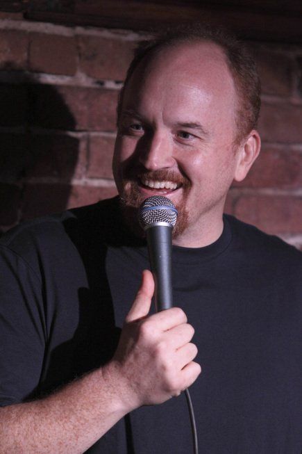 a man holding a microphone in his right hand and smiling at the camera while standing next to a brick wall