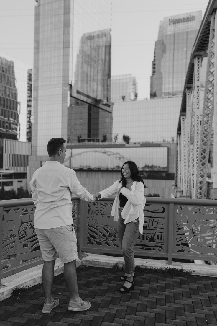 a man and woman are standing on a bridge in front of tall buildings, holding hands