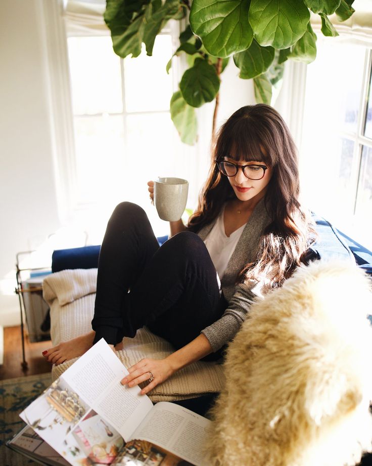 a woman sitting on a couch reading a book and holding a cup in her hand