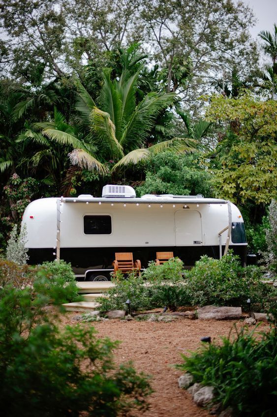 an rv is parked in the middle of some trees and bushes with chairs around it