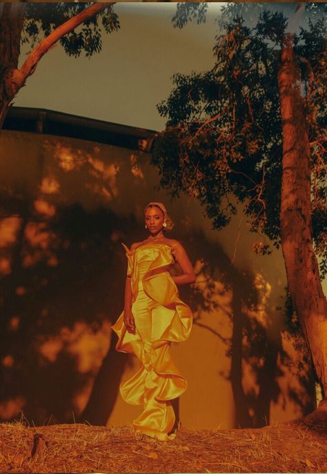 a woman in a yellow dress standing next to trees and a wall with shadows on it