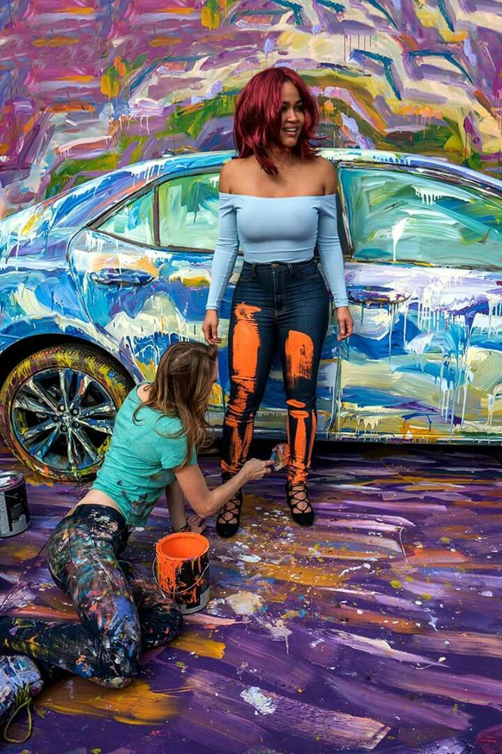 two women are painting a car in front of a colorful wall and another woman is kneeling on the ground