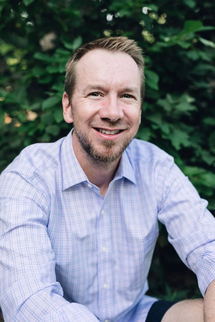 a man sitting on the ground in front of some bushes and trees smiling at the camera