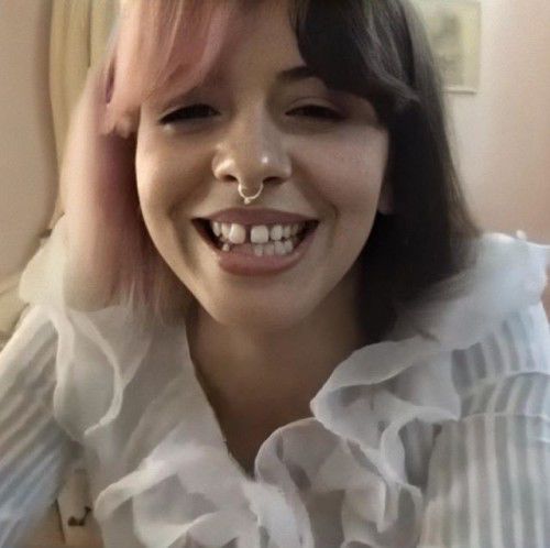 a woman with pink hair and nose piercings smiles at the camera while wearing a white shirt