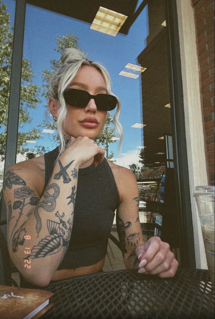 a woman sitting at a table with a drink in front of her and tattoos on her arms