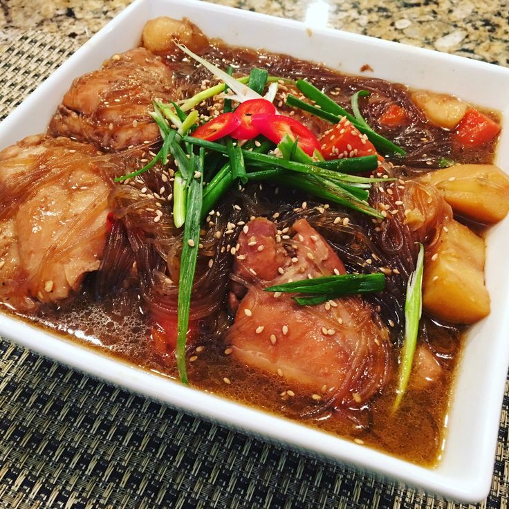 a white square bowl filled with meat and vegetables on top of a table next to a cup of coffee