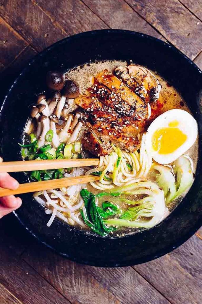 someone holding chopsticks over a bowl of ramen with vegetables and an egg