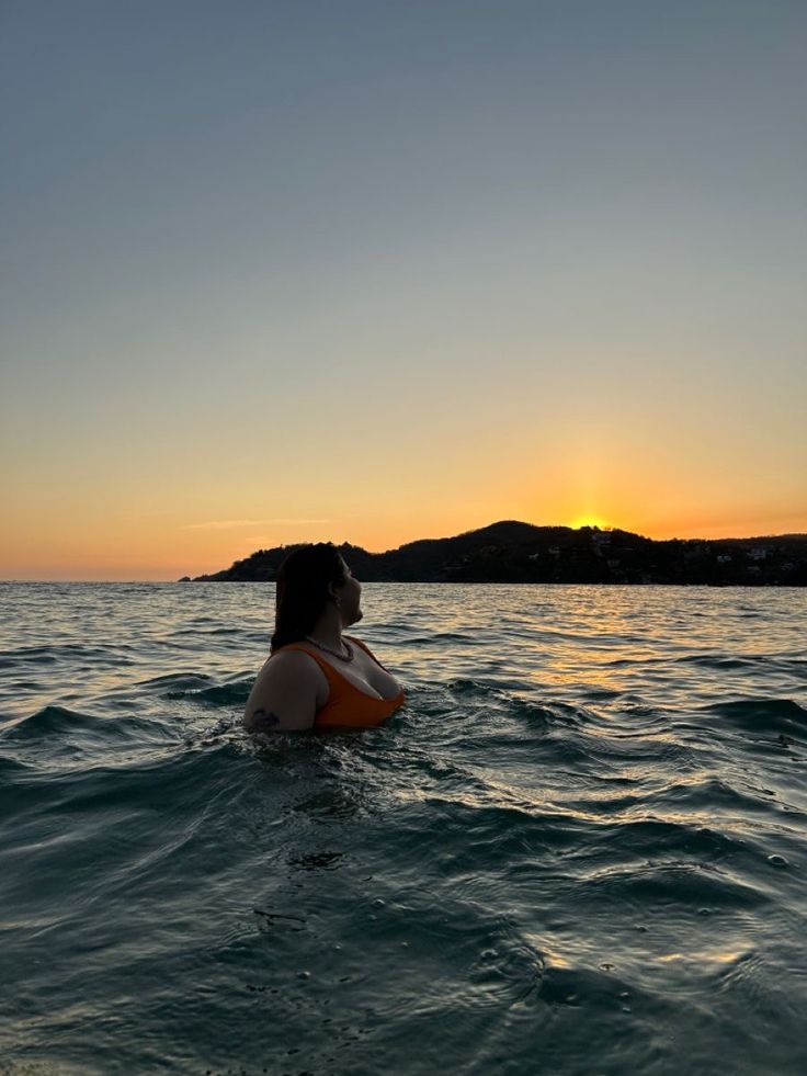 a woman is sitting in the water watching the sun go down on her head and shoulders