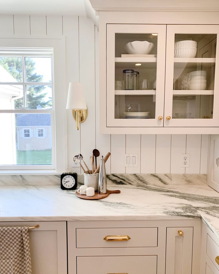 a white kitchen with marble counter tops and gold pulls on the cabinet doors, next to a window