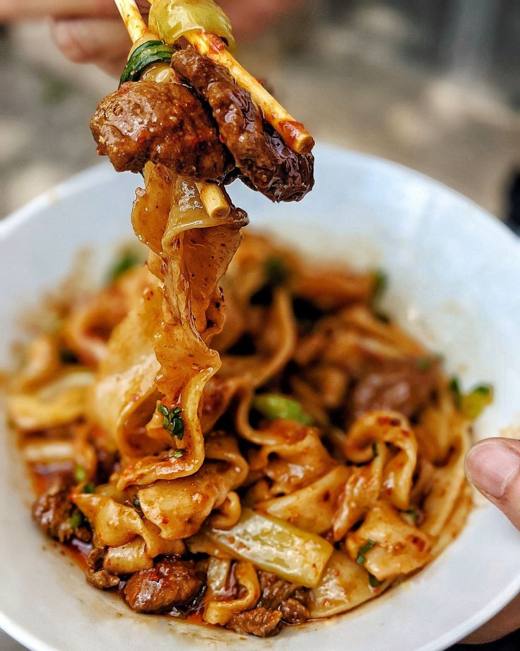 a person holding up a fork full of noodles with meat and vegetables on it in a white bowl