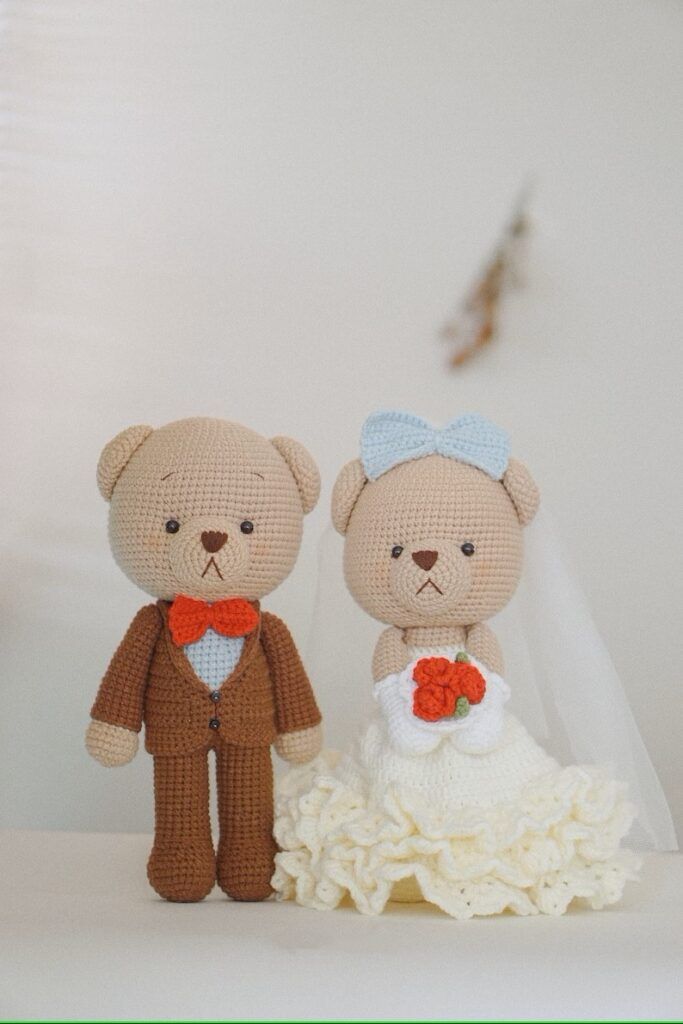 two crocheted teddy bears dressed in wedding attire, one holding a flower and the other wearing a tuxedo