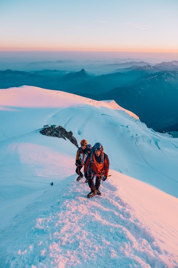 two climbers climbing up the side of a snow covered mountain at sunset or dawn