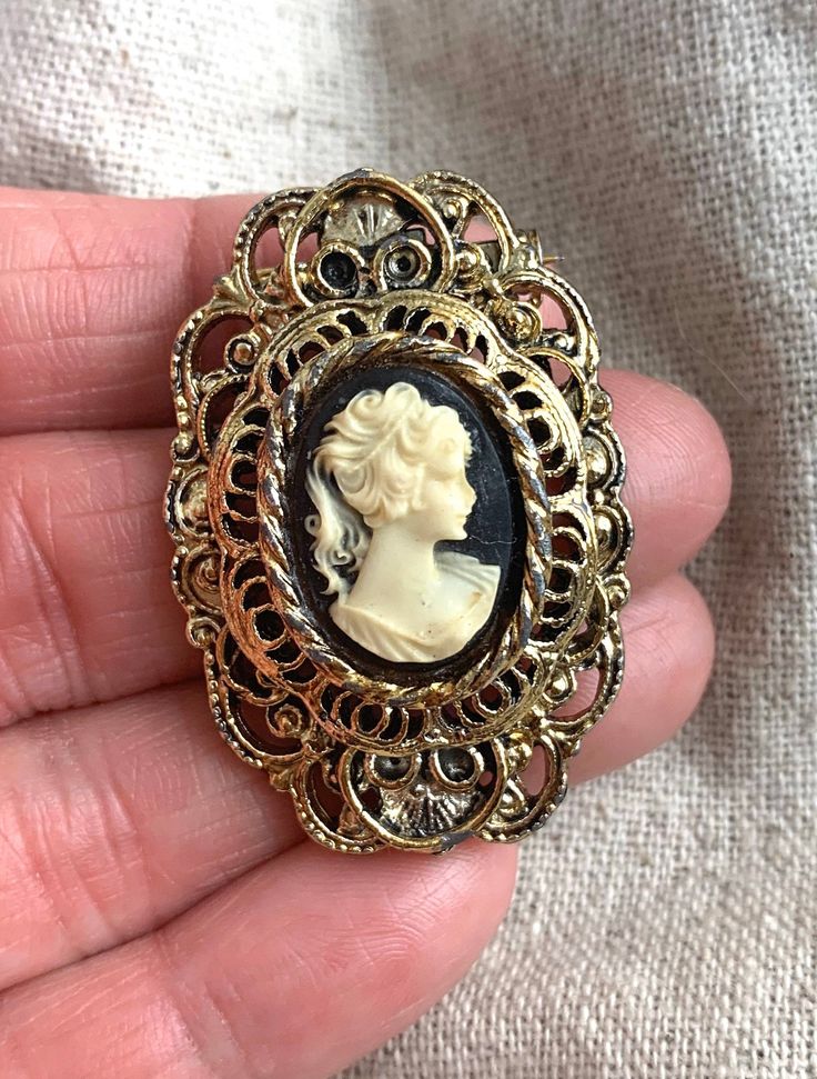 "You will receive this vintage cameo brooch from Gerry's. It measures 1 3/4\" x 1 1/8\" and is cast in an antiqued gold tone base metal. The cameo backing is black and the silhouette is a cream color. This brooch is in very good vintage condition with minimal signs of wear and a properly functioning safety catch. For finished vintage assemblage and vintage inspired necklaces, bracelets, and earrings, please visit us at our other Etsy store, William Dalton Design: www.williamdaltondesign.etsy.com Vintage Medallion Brooch For Wedding, Vintage Medallion Brooches For Wedding, Vintage Brooch With Antique Finish, Antique Cameo Medallion Brooch, Antique Cameo Medallion Brooches, Antique Gold Cameo Brooch, Antique Gold Cameo Brooches, Vintage Medallion Brooches For Formal Occasion, Ornate Medallion Brooches For Collectibles