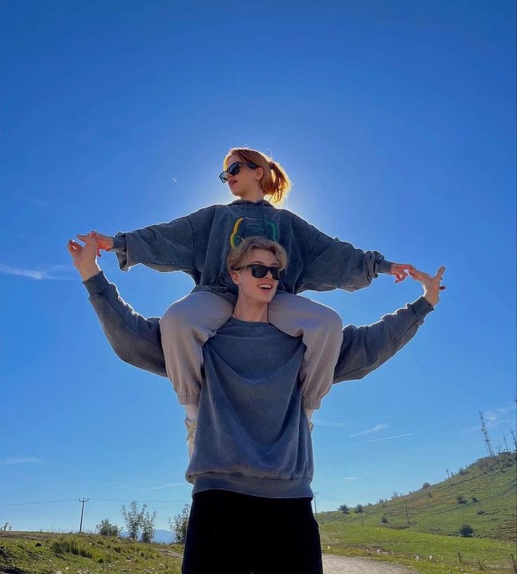 a woman holding onto a man's shoulders while standing on a dirt road in front of a blue sky