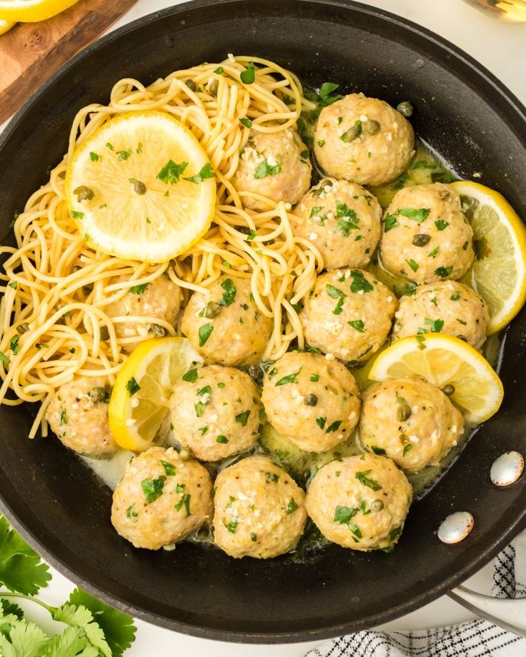 a skillet filled with meatballs, noodles and lemon wedges next to parsley