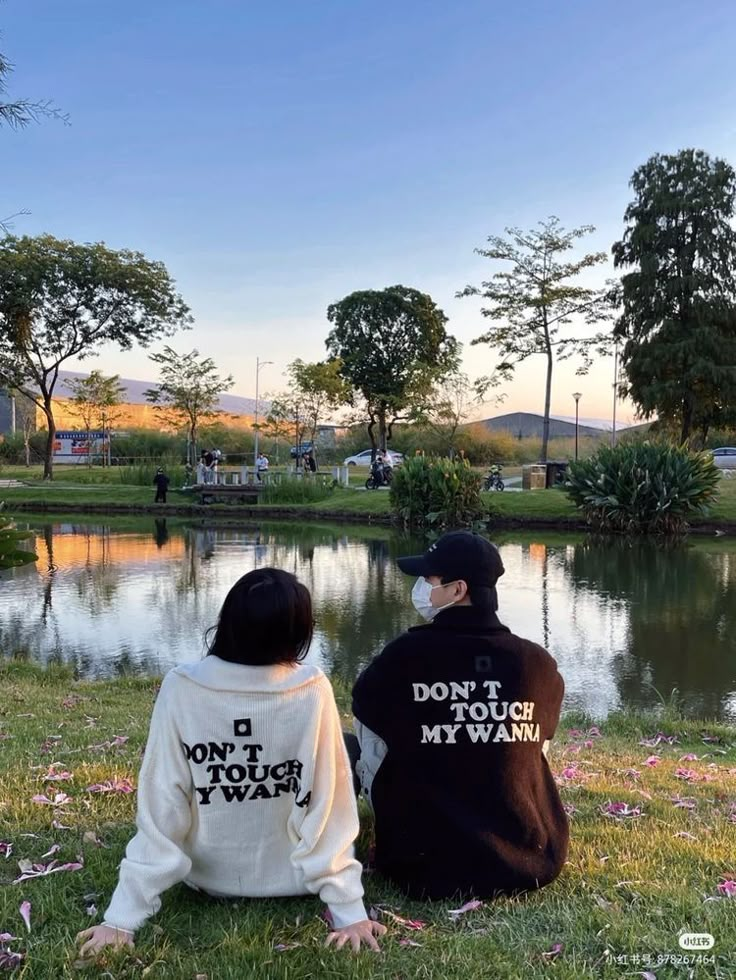 two people sitting next to each other on the grass near a body of water with trees in the background