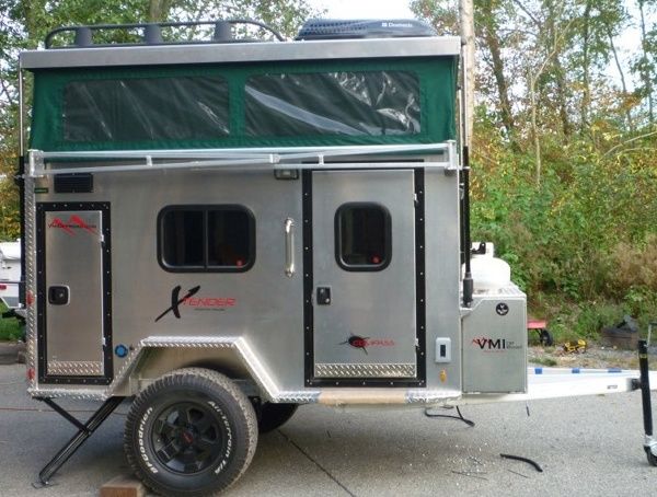 a silver trailer parked on top of a parking lot next to some trees and bushes