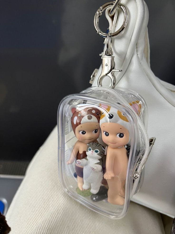 two little dolls are sitting in a clear case on a keychain that is attached to a white purse