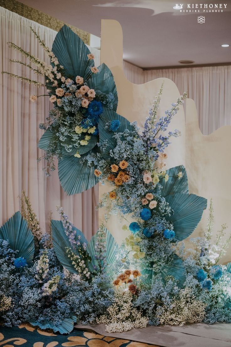 an arrangement of blue flowers and greenery in front of a curtained wall with drapes