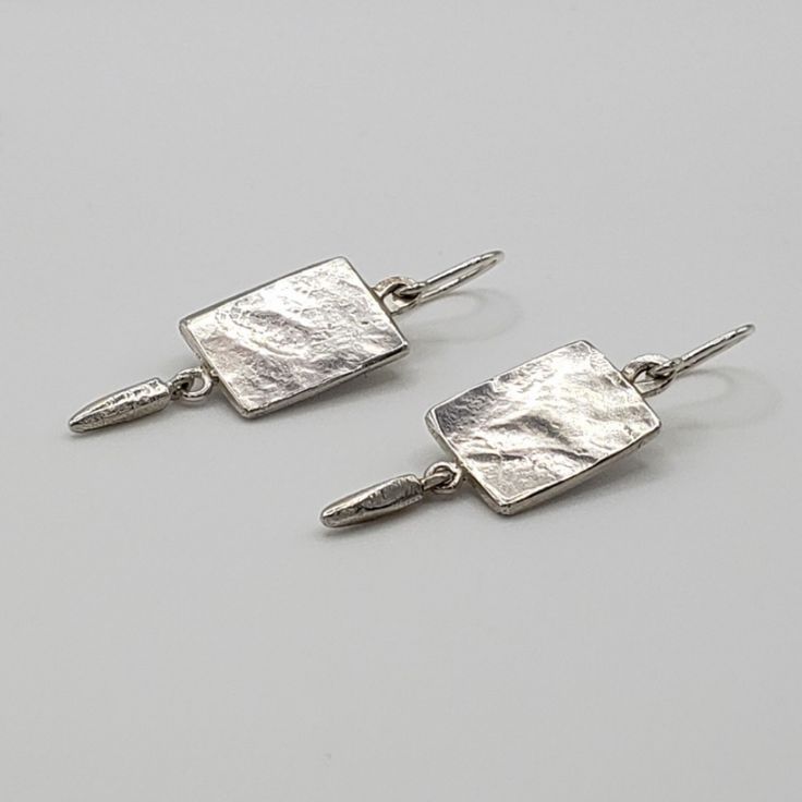 Left facing hammered silver rectangle earrings on gray surface Modern Sterling Silver Hammered Earrings, Modern Hammered Sterling Silver Earrings, Modern Sterling Silver Hammered Linear Earrings, Modern Sterling Silver Linear Earrings With Hammered Detail, Modern Hammered Sterling Silver Linear Earrings, Contemporary Hammered Silver Jewelry, Modern Textured Drop Earrings, Silver Hammered Sterling Silver Linear Earrings, Hammered Sterling Silver Long Drop Earrings