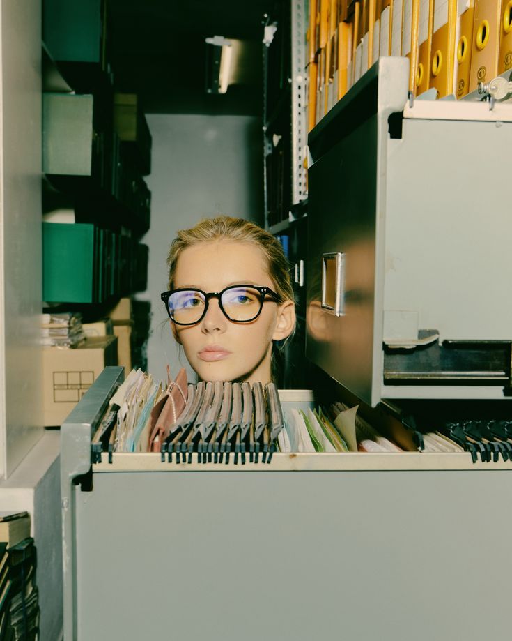 a woman wearing glasses standing in front of a filing cabinet filled with files and folders