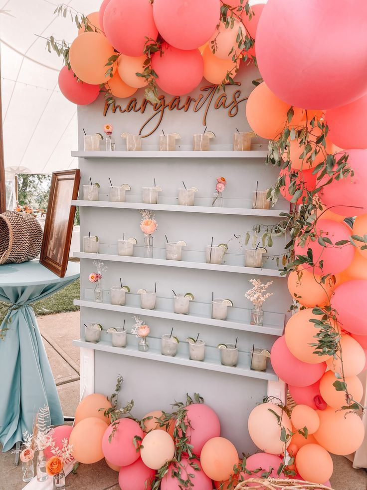 a table topped with lots of pink and orange balloons next to a white shelf filled with candles