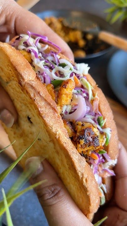 a person holding a sandwich with coleslaw and other food items in the background