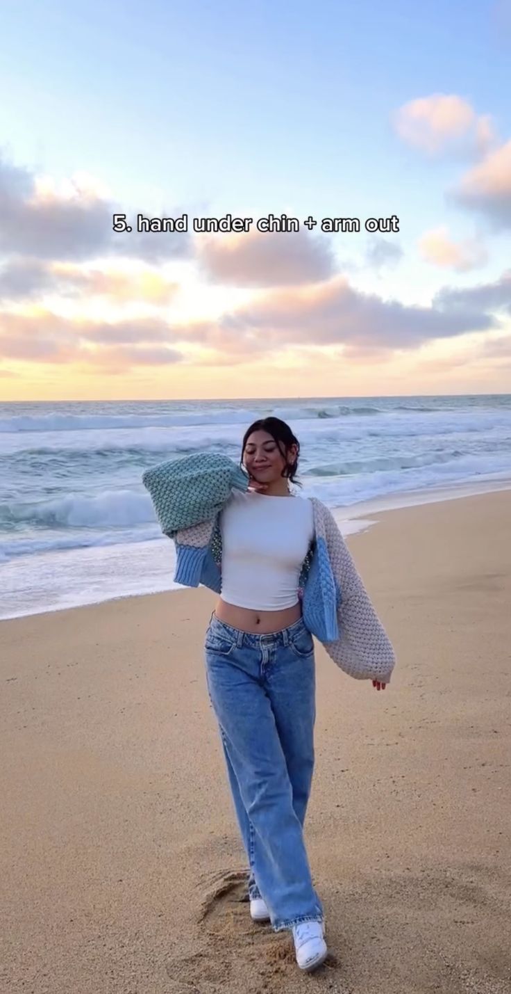 a woman walking on top of a sandy beach next to the ocean with a sweater over her shoulders