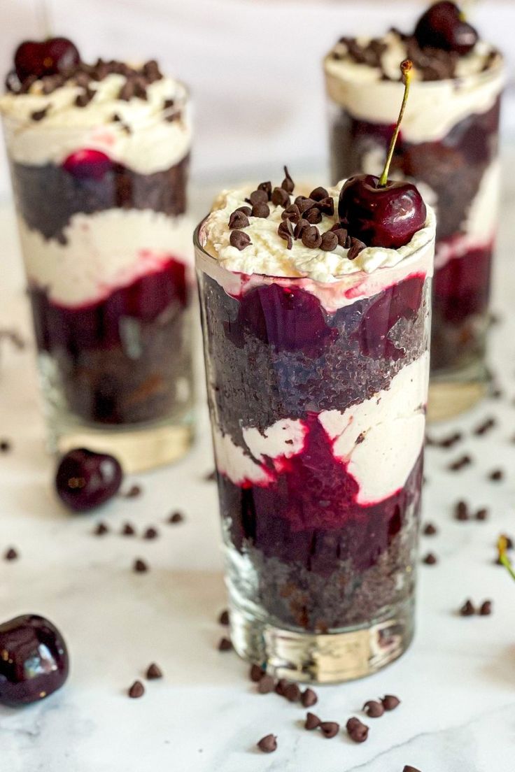 three desserts with cherries and whipped cream in them on a white tablecloth