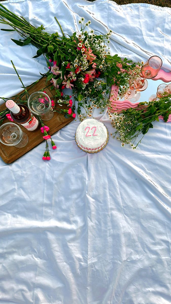 a table topped with flowers and cake on top of a white sheet covered ground next to wine glasses