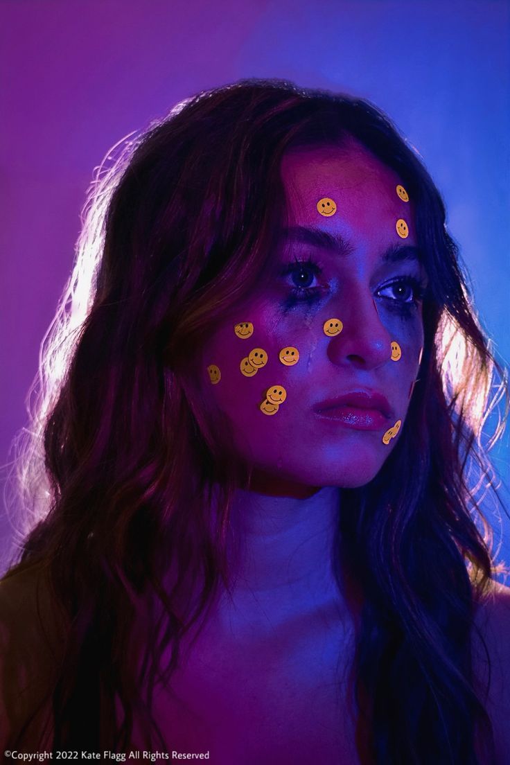 a woman with yellow smiley faces painted on her face in front of a purple background