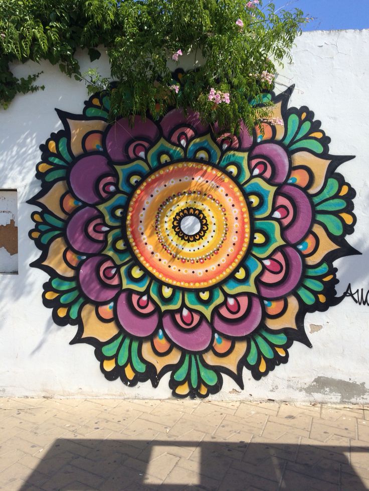 a large colorful flower painted on the side of a building next to a tree and sidewalk