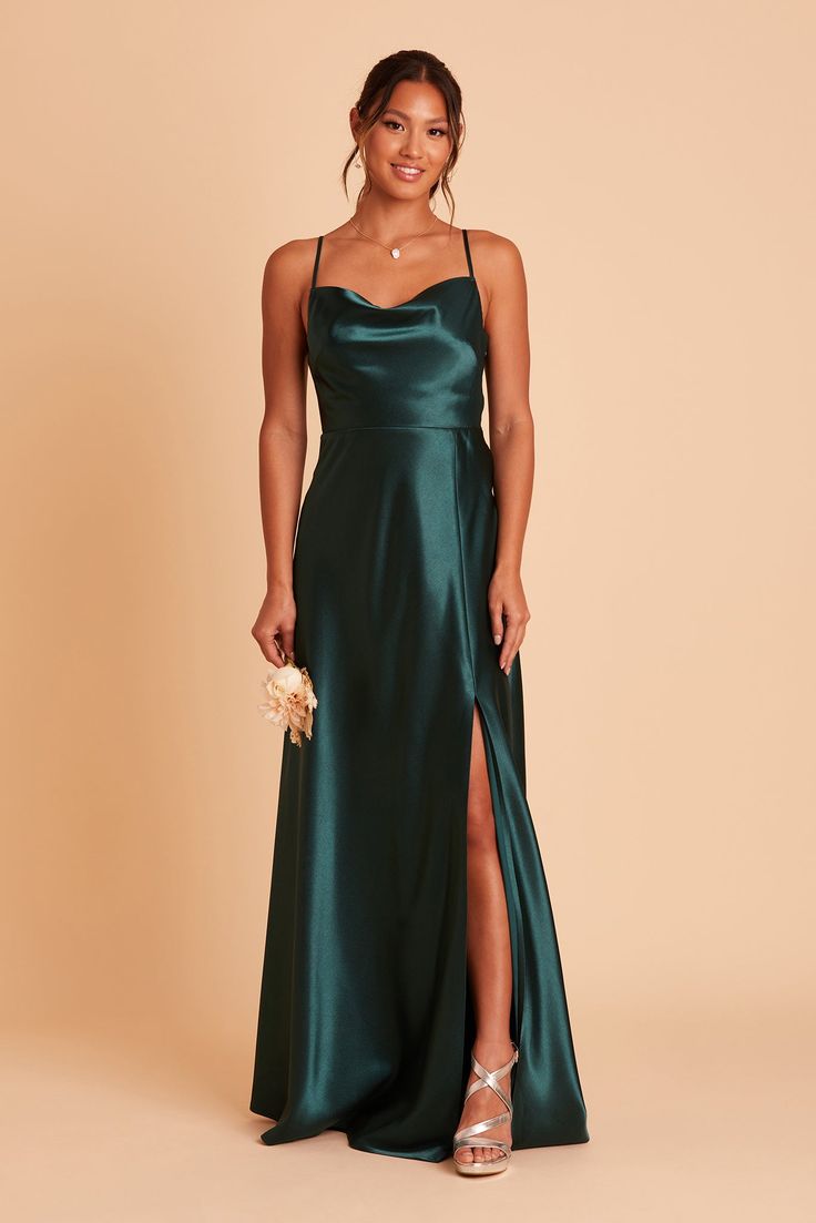 a woman wearing a green satin gown with side slit and spaghetti neckline, standing in front of a beige background