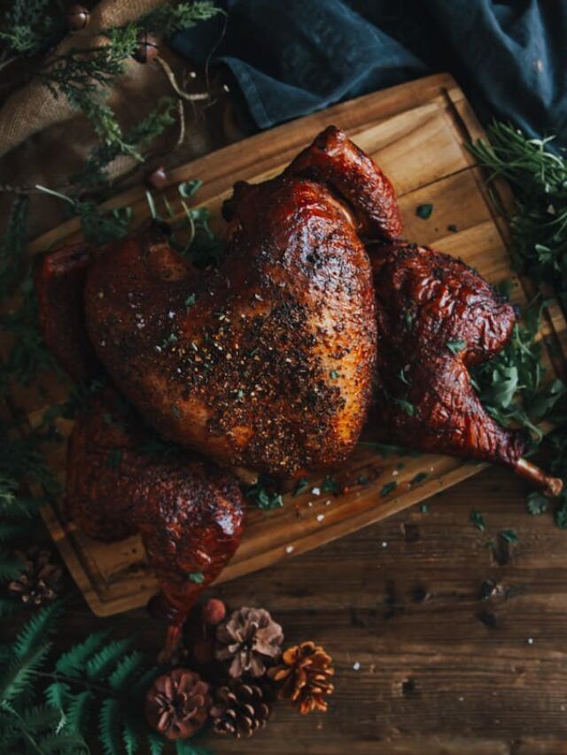 two roasted turkeys on a wooden cutting board with pine cones and evergreen leaves around them