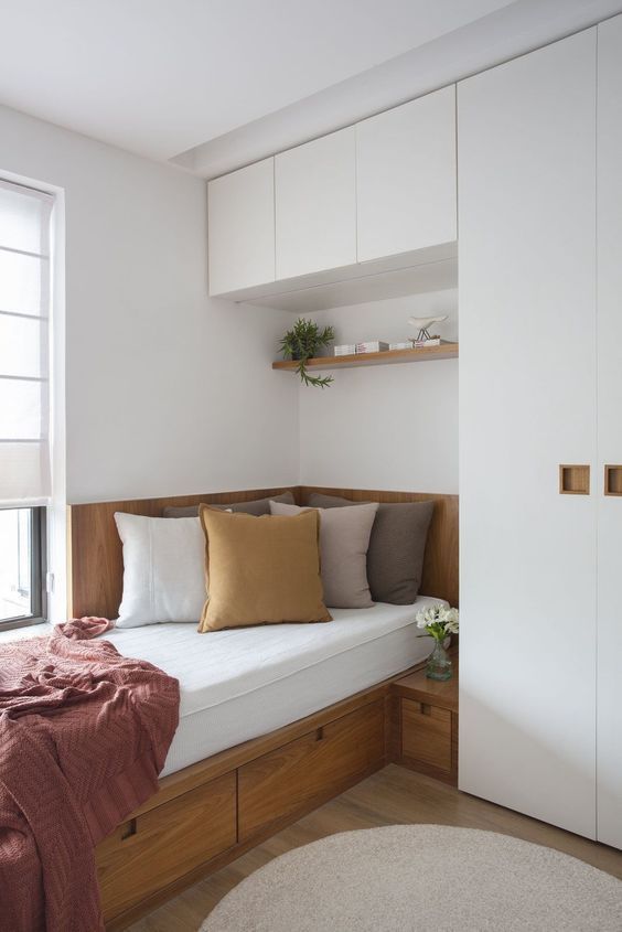 a bed sitting under a window next to a white cupboard filled with lots of pillows