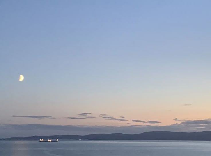 a full moon is seen over the ocean with boats in the water and mountains in the distance