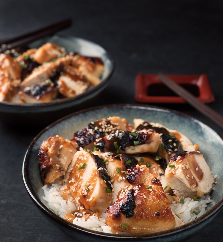 two plates filled with chicken and rice on top of a table next to chopsticks