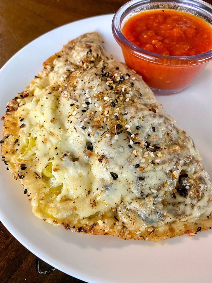 a white plate topped with an omelet covered in cheese next to a bowl of sauce