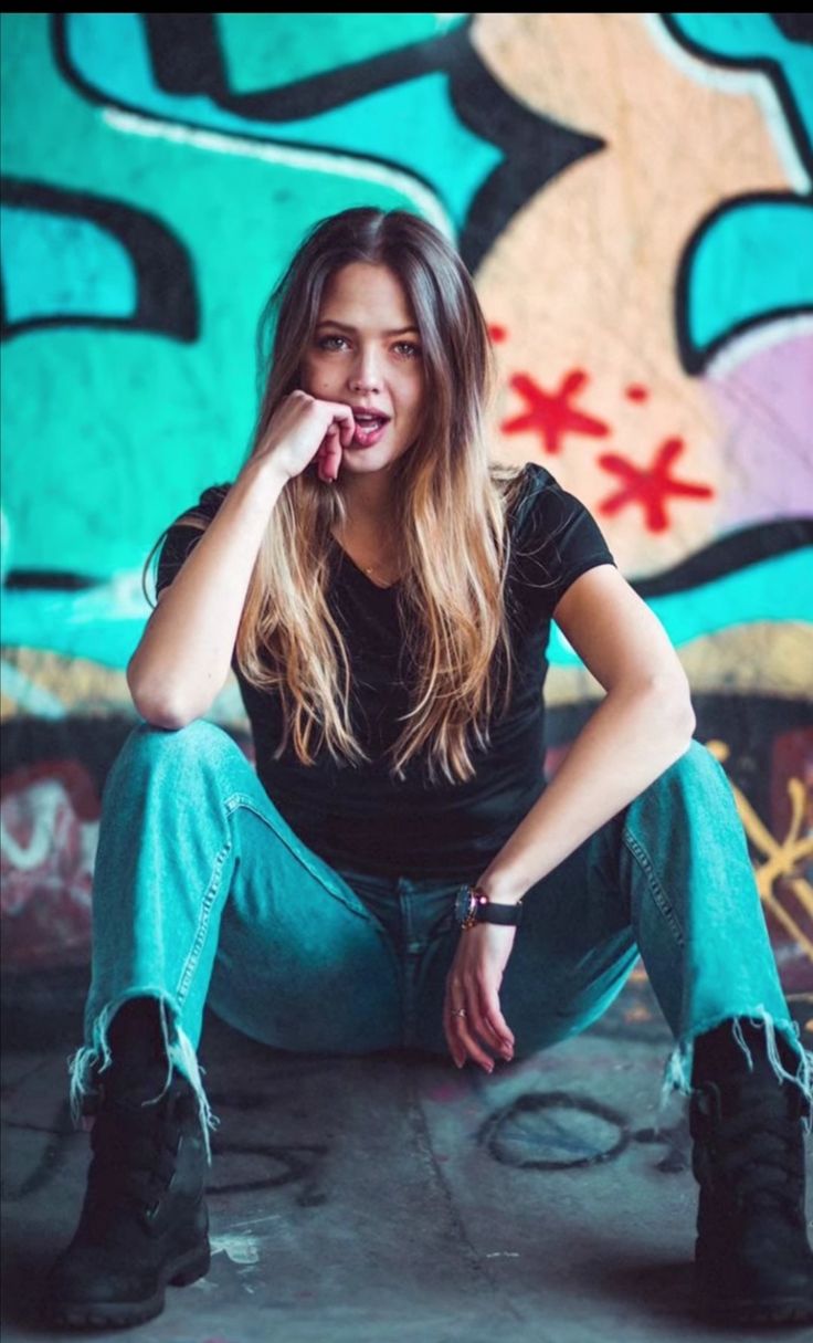 a woman sitting on the ground in front of a graffiti covered wall with her hand under her chin