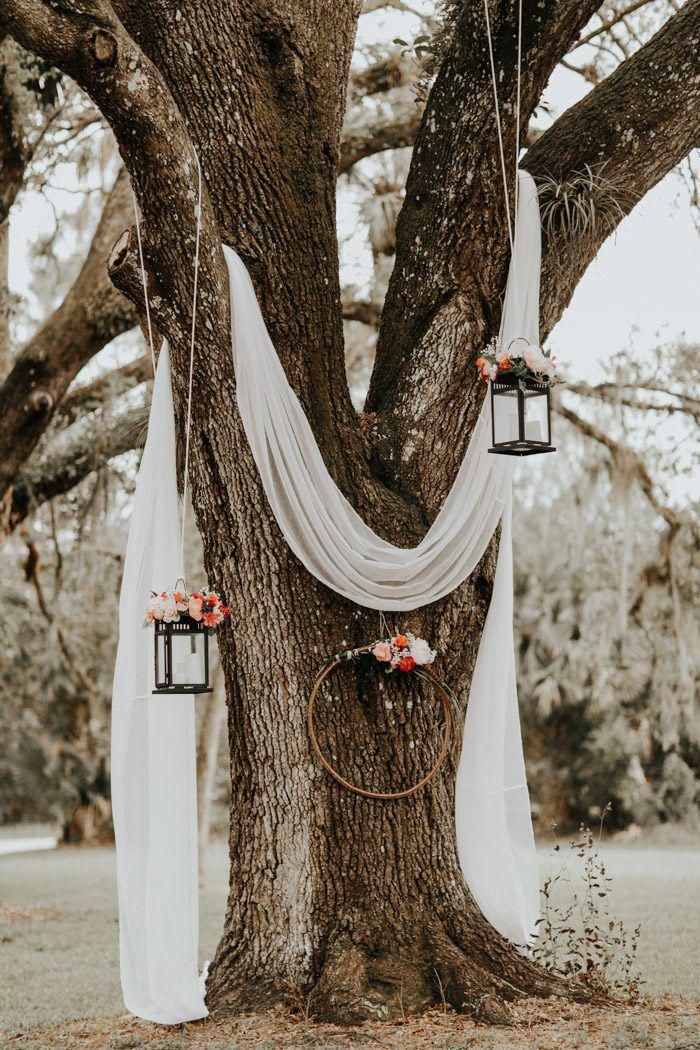 an outdoor wedding ceremony with hanging lanterns and draping on the tree trunk, decorated with flowers