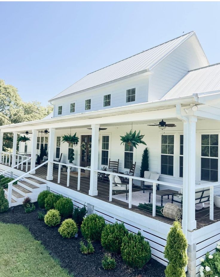 a white house with porches and chairs on the front lawn, surrounded by greenery