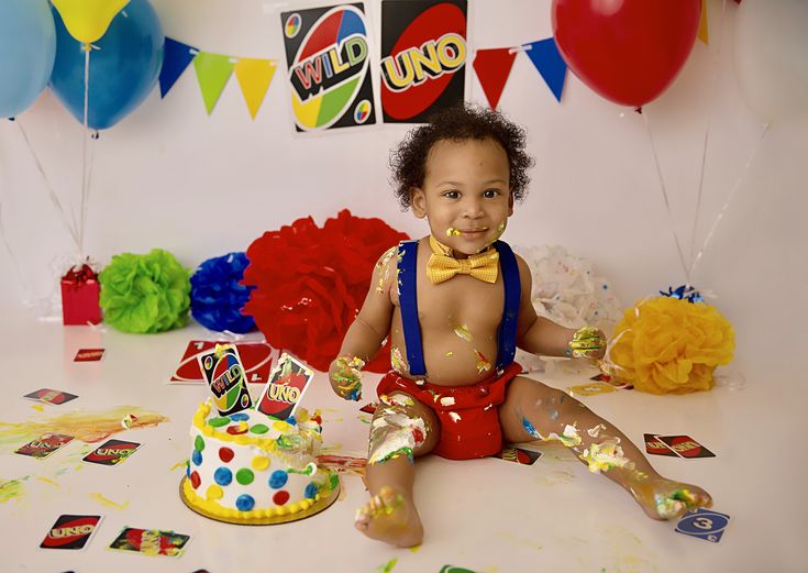 a baby sitting on the floor in front of a birthday cake and balloons with confetti all over it