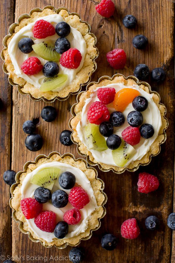 three small pies with fruit on top are sitting on a wooden table next to berries and kiwi