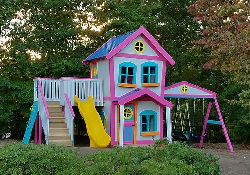 a pink and blue house with a yellow slide in the front yard next to some bushes