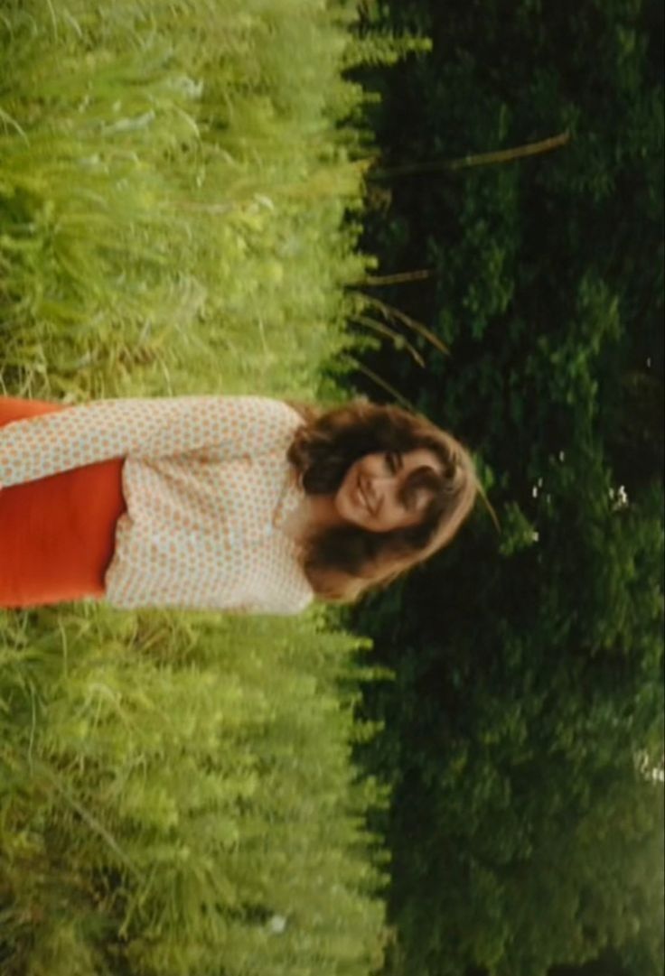 a woman standing next to a lush green forest filled with lots of tall grass and trees